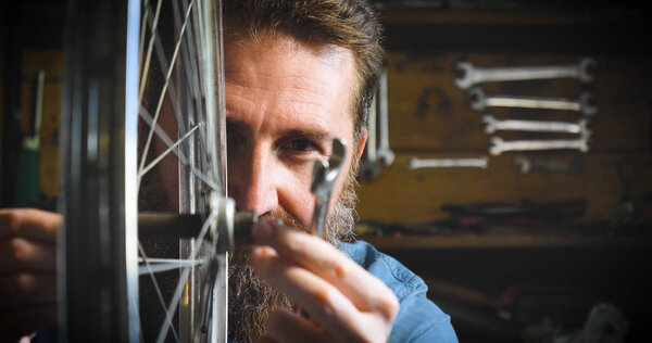 Well-groomed handsome bearded master hipster, specialist in bicycles, repairing a bicycle in his workshop, wheels, frame, spokes, the background of tools. Concept: pro bike, cycle passion, lifestyle.