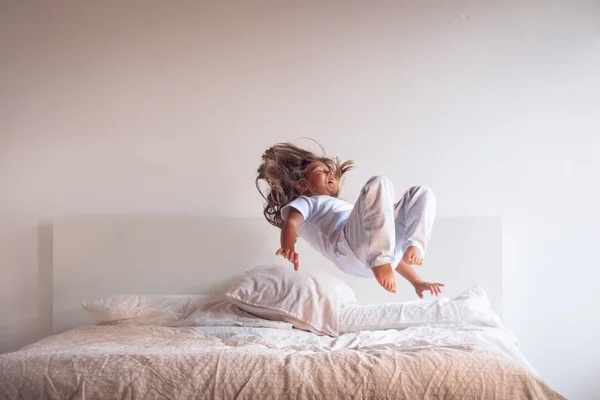 A little girl, wearing white pajamas, jumps on the bed and has fun. Concept of youth, wellness and health, happiness and freedom.