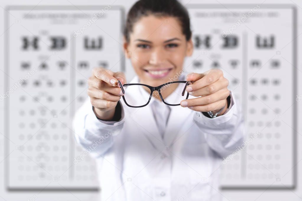 happy smiling woman in optical center standing at Snellen chart used for visual acuity testing and giving glasses 