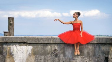 Yemyeşil bir kırmızı elbiseli güzel bir ışık balerin portresi bir taş iskele, ihale oturur. Konsept: balet, ince, diyet, hafif kadın, esneklik, güzel rakam, sağlıklı yaşam, gökyüzü.