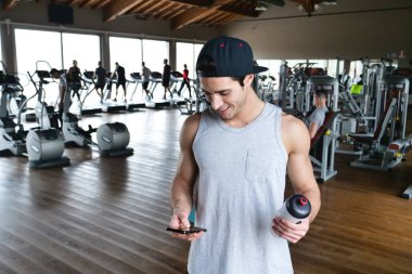Bir egzersiz ve diğer arasında istirahat ederken bir spor salonunda bir adam portresi telefonunu sosyal ağlar izlemek veya iletileri göndermek için kullanır. Kavramı: spor, kişisel antrenör, iletileri, telefon
