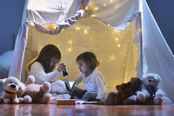 二人の少女は 懐中電灯で本を読むと 中に眠るキャンプ テントを構築する夕方自宅プレイします ゲーム マジック 創造性 警報システム — ストック写真