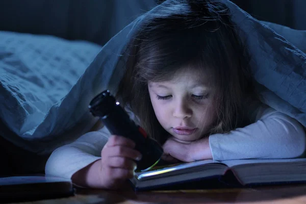 A little girl at night under the covers reads a book of adventures and fantasy and holds a torch in her hands to make herself light. Concept of: educational, magic, creativity, adventure and fantasy.