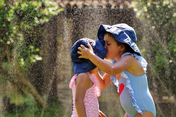 Two little girls play in the garden with water, the two girls run, get wet and laugh. Concept of: family, love, fun and happiness.