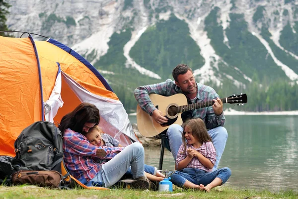 A happy family camping at the lake, playing the guitar and singing a song together in front of the tent. Concept of: trekking, nature, family.
