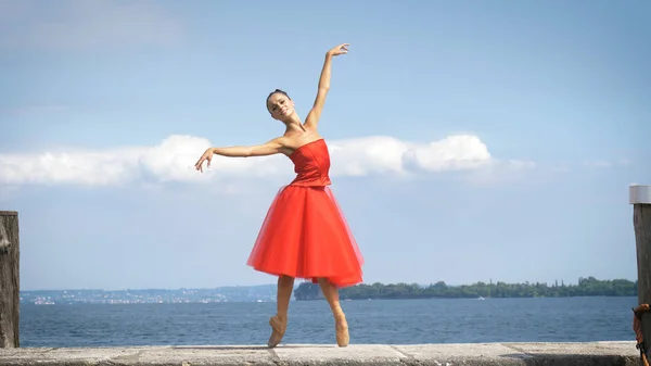 一个美丽的浅色芭蕾演员的肖像 穿着郁郁葱葱的红色礼服 背景是湖和天空 芭蕾舞演员 轻盈的女人 美丽的身材 健康的生活方式 — 图库照片