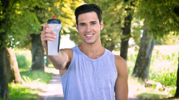 肖像画英俊的年轻人运动员显示水瓶 而站在绿色公园户外 — 图库照片