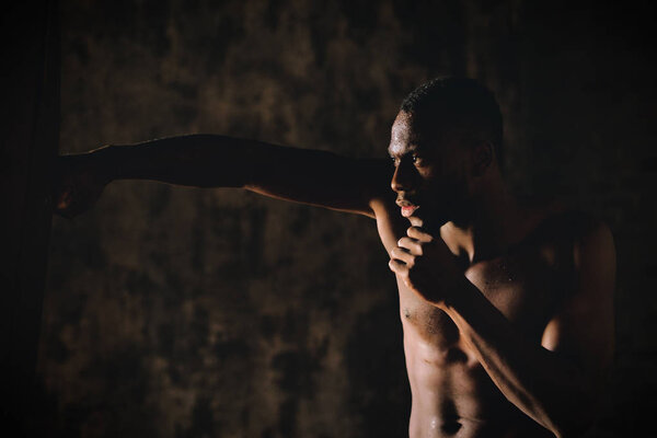 handsome shirtless muscular african man boxing punching bag in dark room 