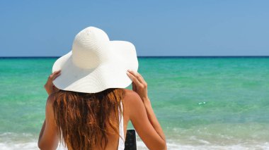 Güzel kadın denizde bir beyaz mayo içinde beyaz bir şapka arkası deniz mavi su arka bilgisayar çalışma kum üzerine oturuyor.