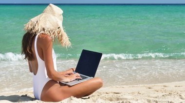 Çalıştığını onun bilgisayar denizin, arkası beyaz mayo, bir arka plan mavi deniz suyu giyen bir hasır şapka giyen güzel bir kadın kum üzerine oturur.