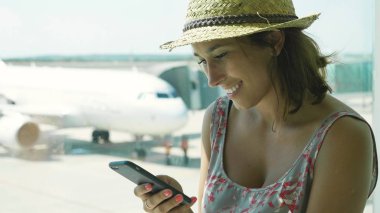 Gülümseyen, uçak telefon, bir elbise, hasır şapka, mesaj yazarken onu uçuş için bekleyen bir genç turist kız havaalanında, portresi. Konsept: dünyayı, tatil, tatil.