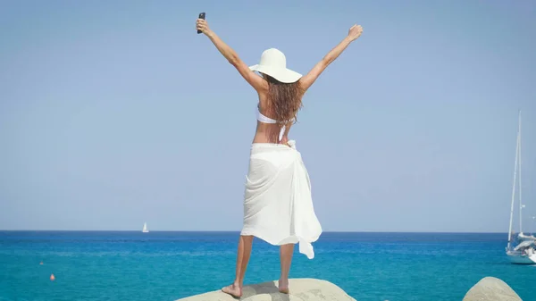 A beautiful young woman smiling, feels happy, in her hand a phone, in a white bikini, in a hat, sunglasses, a background of sea blue water and yachts. Concept: sea rest, sun, travel, vacation, freedom