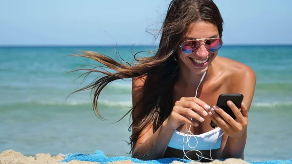 Beautiful girl laughs happily and listens to music on the phone at the sea wearing sunglasses, in a white bathing suit, background of sea blue water.