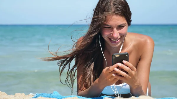 Beautiful girl laughs happily and listens to music on the phone at the sea wearing sunglasses, in a white bathing suit, background of sea blue water.