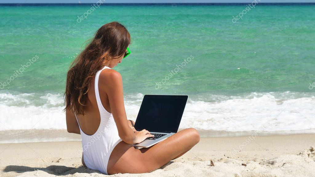 A beautiful woman sits in the sand, working in the laptop at the sea, wearing sunglasses, wearing a white bathing suit, a background of sea blue water.