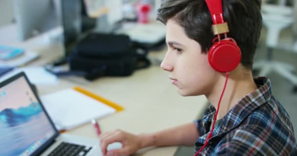 年轻学生男孩在桌子上学习 使用笔记本电脑和戴红色耳机的视频 — 图库视频影像