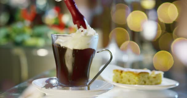 咖啡馆里的热巧克力圣诞节的时候 一个酒保在一个透明的杯子里喷上一些新鲜的奶油 热巧克力烟和圣诞灯在背景 温暖和节日 — 图库视频影像
