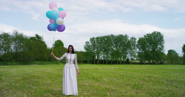 カラフルな風船を押しながら芝生の上に立って長い白いドレスを着た女性のスロー モーション ビデオ — ストック動画