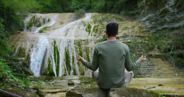 林や岩の上に座って ヨガ瞑想の滝で旅行者の男の動画 — ストック動画