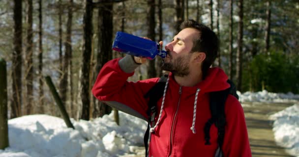 ボトルから飲むハイカー旅行者の男性が冬の森で身に着けている赤いジャケットとバックパックのビデオ — ストック動画