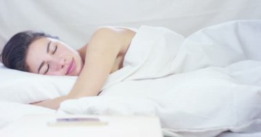 Güzel bir genç kız sıcak bir yatakta zayıf up için çalar saat kapalı geçiş için basit bir çalışma günü kaplı bir yumuşak sıcak beyaz battaniye, beyaz bir arka plan üzerinde ile.