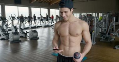 Bir egzersiz ve diğer arasında istirahat ederken bir spor salonunda bir adam portresi telefonunu sosyal ağlar izlemek veya iletileri göndermek için kullanır. Kavramı: spor, kişisel antrenör, iletileri, telefon