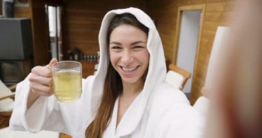 Güzel bir kadın, beyaz bornoz giymiş bir sağlık merkezinde sıcak bir bitki çayı içer ve spa göstermek için video çağrısı yapar. Kavramı: teknoloji, sakin ol, sosyal ağ.