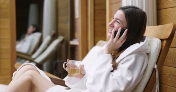 白いバスローブに身を包んだ美しい女性は 熱いお茶を飲むし 彼女の電話を使用してメッセージを送信します リラックスして ネットワーク — ストック動画