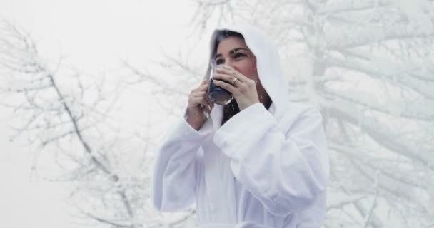 白いバスローブで美しい女性は 雪が空から落ちている間山で熱いお茶を飲みます リラクゼーション — ストック動画