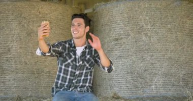 Yakışıklı genç bir çiftçi (öğrenci), bir gömlek bir mesaj, selfie, yazma Evet bir haystack üzerinde oturuyor mutlu gülümseyerek. Konsept: iletişim, ekoloji, tarım, saman, kişiler.