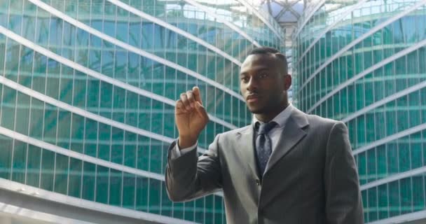 指ホログラム金融チャートに触れ 現代のガラスの建物に立ってスーツを着たアフリカ系アメリカ人のビジネスマン ビデオ — ストック動画
