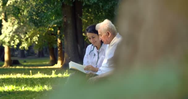 Video Vårdgivare Sjuksköterska Med Pensionärs Man Läsning Bok Park Bänken — Stockvideo