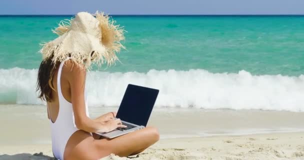 慢动作视频的妇女在帽子坐在沙滩上 并使用笔记本电脑 — 图库视频影像