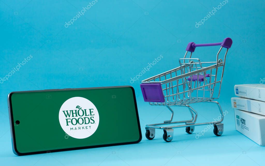 Dhaka, Bangladesh- 03 July 2025: Smartphone with Whole Foods Market logo next to a small shopping cart and boxes against a blue background.