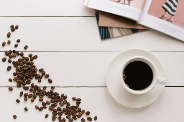 Xícara de café preto em uma mesa de madeira branca com grãos de café e revista de moda. Processamento de autor, efeito de filme . — Fotografia de Stock