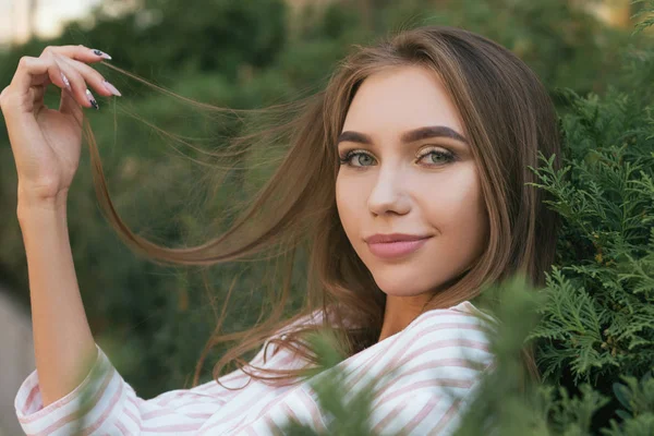 Joven chica atractiva cerca del arbusto verde, tocando para el cabello. Chica sonriente con cabello largo, imagen con efecto de película y procesamiento de autor . —  Fotos de Stock
