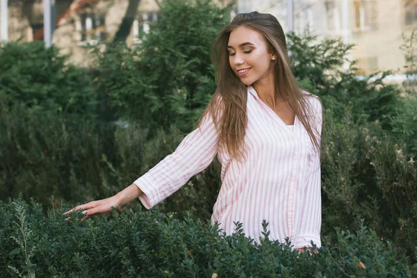 Menina bonita nova na cidade, frente do arbusto verde estão tocando ramos. Menina sorridente com cabelos longos, imagem com efeito de filme e processamento de autor . Imagem De Stock