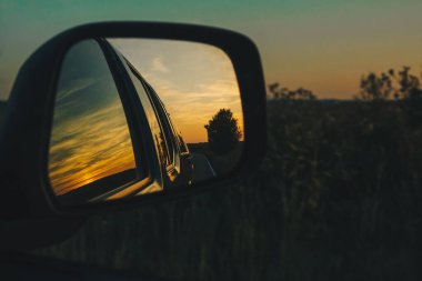Gün batımında ve alacakaranlıkta çayırın önündeki dikiz aynasından yola bak. Arabadan tarlaya güzel bir manzara. Seyahat konsepti, tatillerde maceralar.