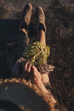 Beautiful young woman is sitting in the meadow with a brunch of hydrangea in her hand. End of summer, girl is happy and enjoying warm sunny day. Author color correction and stylish outfit matching to autumn colors.