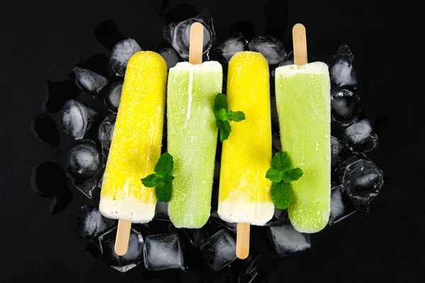 Yellow and green fruit ice cream on a stick with mint on ice on a black table