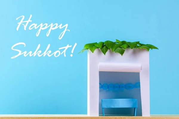 Mutlu Sukkot metni. Kağıttan yapılmış bir kulübe mavi bir arka plan üzerinde yapraklarla kaplı. Kartpostal, tebrikler
