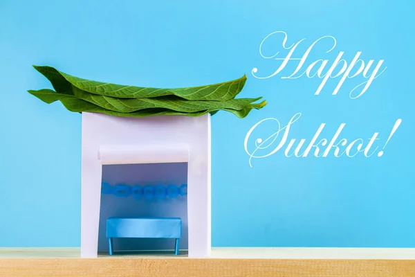 幸せな仮庵の祭りのテキストです 紙で作られた小屋は青い背景の葉で覆われています はがき お祝いの言葉 — ストック写真