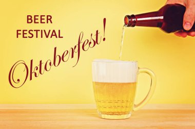 Bir adam bira kupa ahşap bir masa üzerinde sarı bir arka plan üzerine bir şişe içine dökülen. Octoberfest