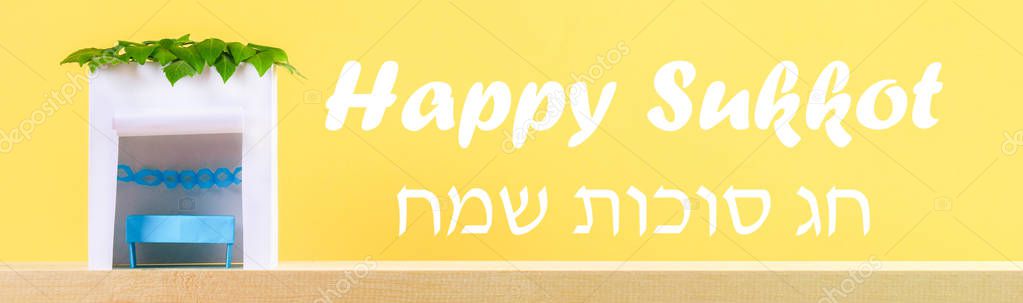 Banner. The text in Hebrew is Happy Sukkot. A hut made of paper covered with leaves on a yellow background