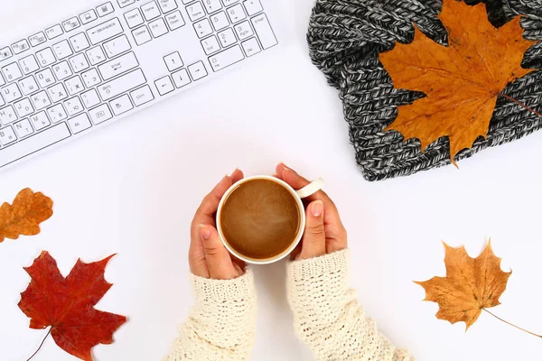 Coffee with marshmallow in hands on a white desktop next to an empty block and keyboard, autumn leaves. Fall mood. Top view, flat layout. Copy the space