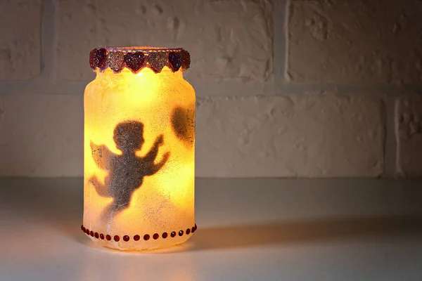 Beyaz tuğlalı arka planda Diy Peri Kavanozu. Hediye fikirleri, dekorasyon St. Şubat 14, Sevgililer Günü, aşk, düğün. El yapımı lamba, gece lambası, cam kavanozdan fener, peçete, yapıştırıcı, sim.