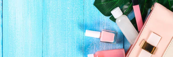 Cosmetics in a pink cosmetic bag. Lip gloss, cream, nail polish, skin care products on a tropical leaf on a blue wooden background