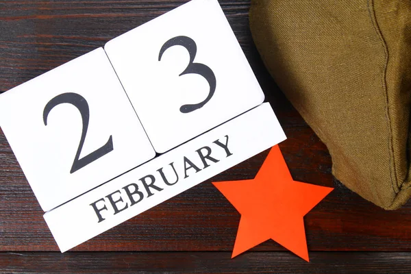 White wooden perpetual calendar with the date of February 23 on a wooden table. Defender of the Fatherland Day. Red star and military cap. Top view.