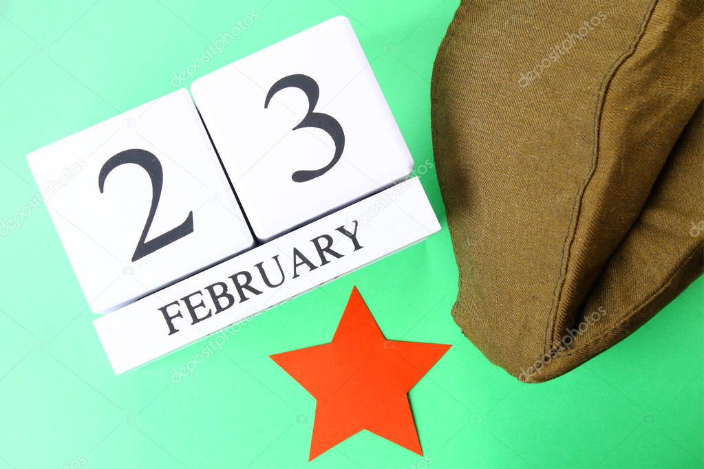 White wooden perpetual calendar with the date of February 23 on a green background. Defender of the Fatherland Day. Military cap and red star. Top view.
