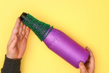 DIY Mardi Gras şişe mor kendinden yapışkanlı kağıt, yeşil boncuk, karnaval maskesi, payetler sarı arka plan. Hediye fikri, dekor Mardi Gras, yağ, Shrove Salı. Adım adım. Üstten görünüm. İşlem çocuk zanaat